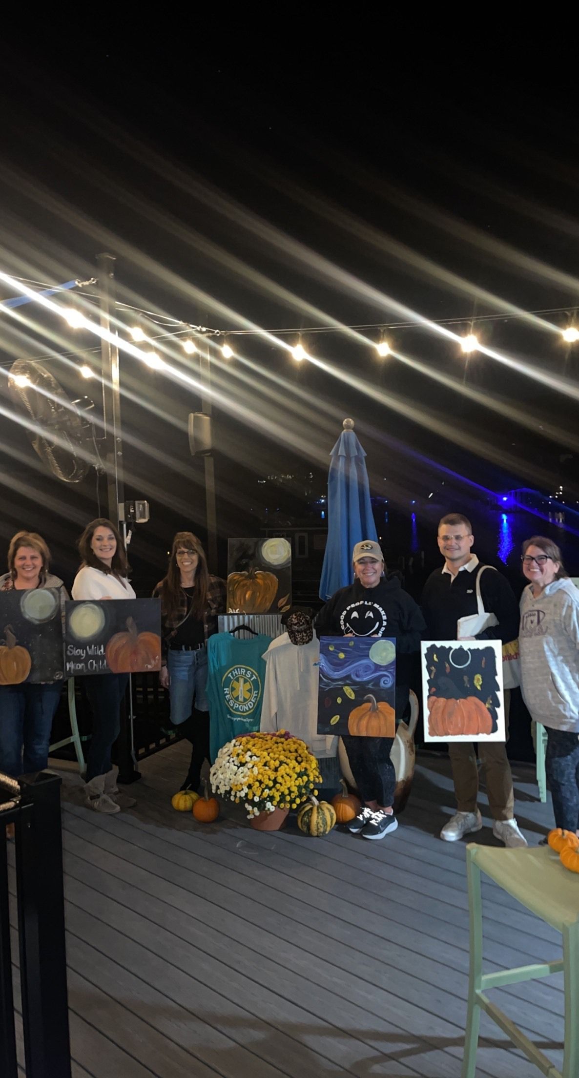 Group posing outdoors at night under string lights, holding flowers and signs near a memorial.