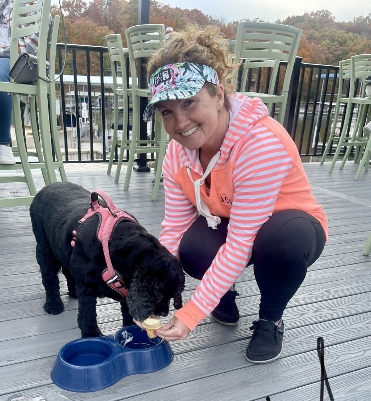 Woman kneels beside a black dog drinking from a blue bowl on a wooden deck.