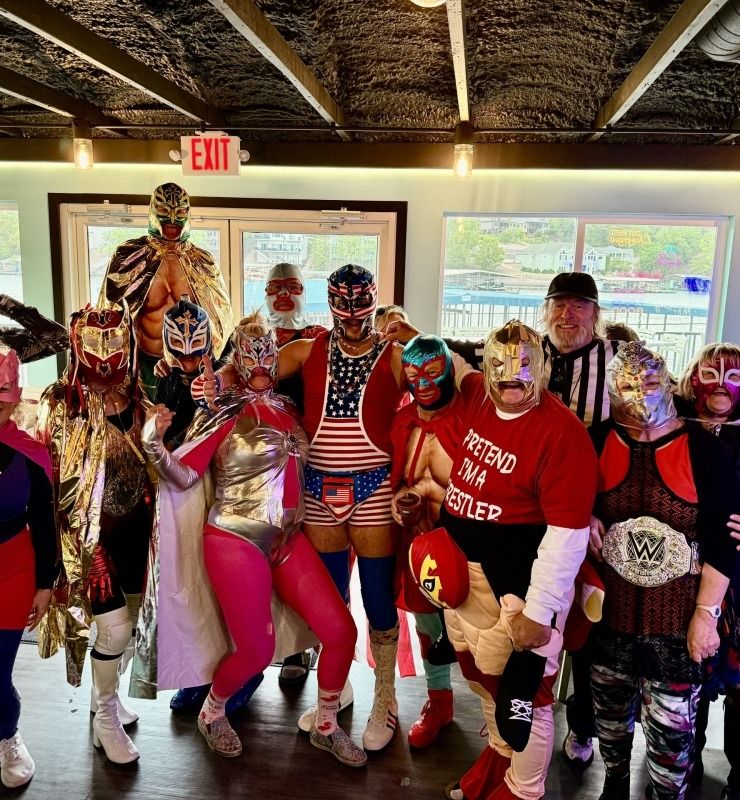Group of people in colorful costumes posing indoors, with window light and an EXIT sign behind them.