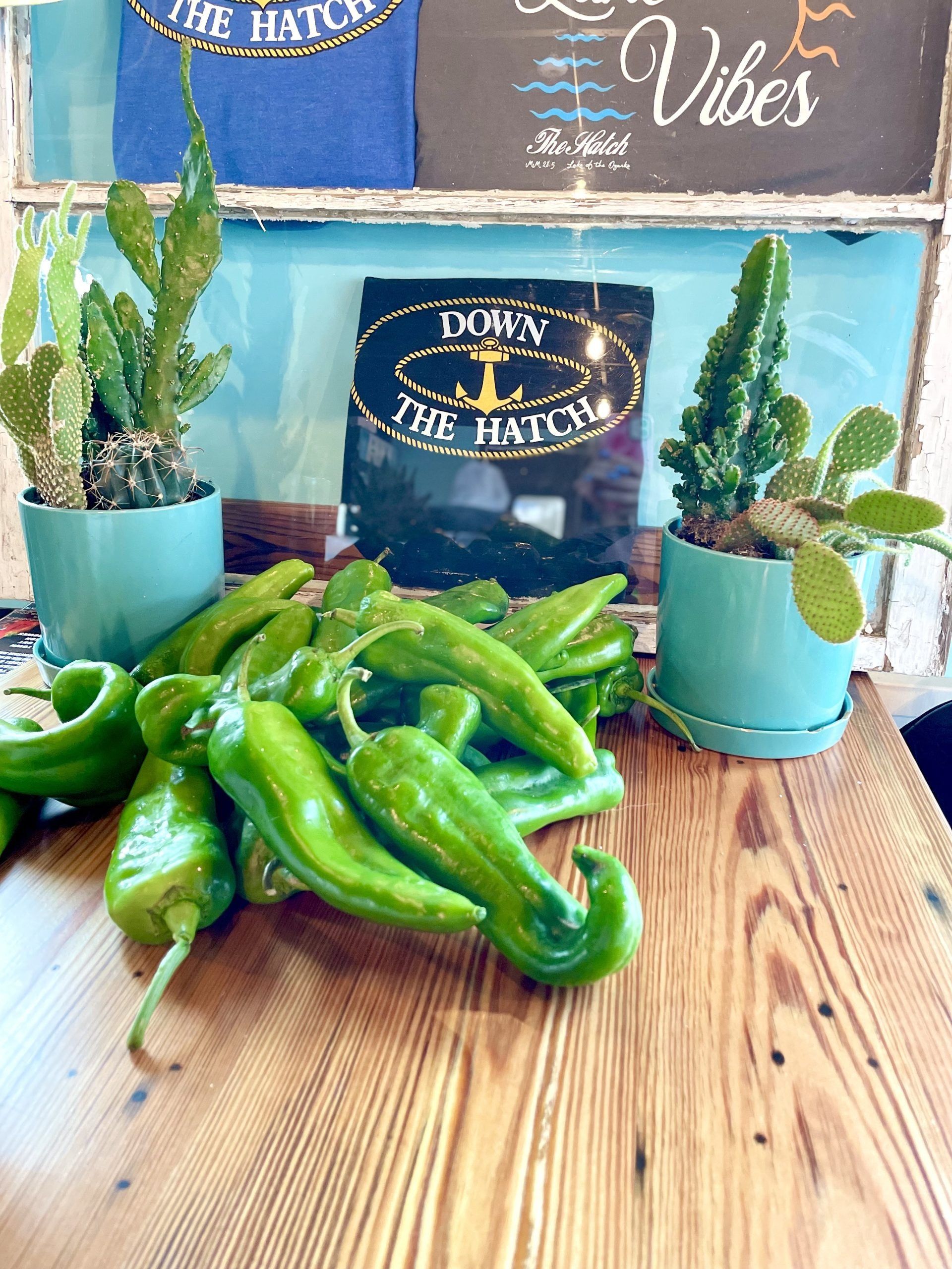 Pile of green peppers on a wooden table with potted plants and a sign in the background