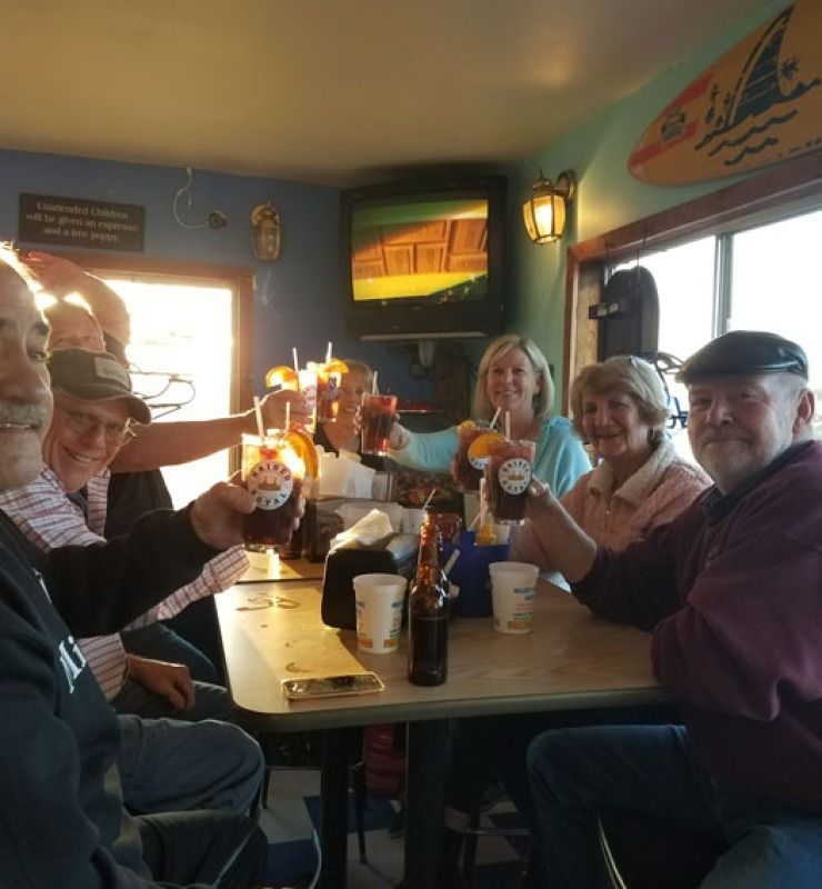 Group of people at a restaurant table raising drinks and smiling, with warm lights and a TV in the background