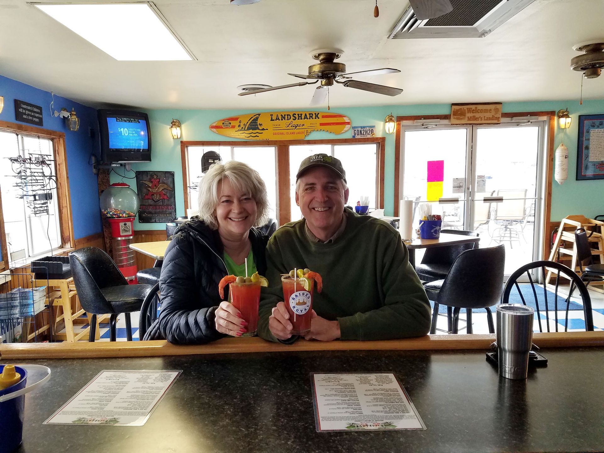 Two people smiling behind a bar, holding colorful drinks in a casual cafe or pub.