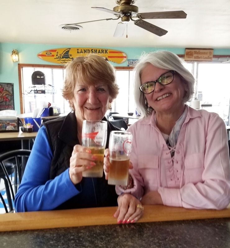 Two women smiling and toasting drinks at a bar with a surfboard sign overhead