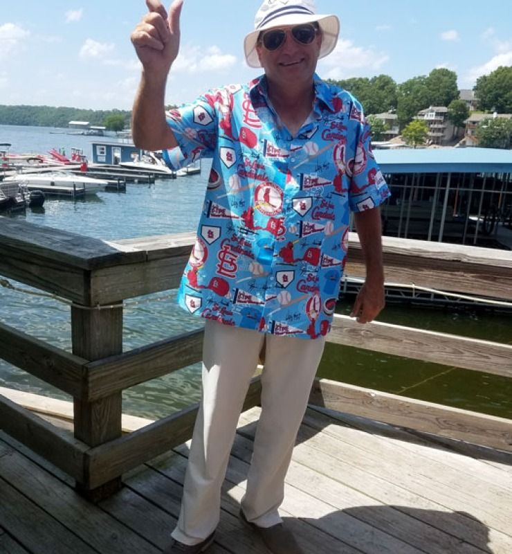 Man in a blue patterned shirt and hat standing on a dock, pointing upward by the water.