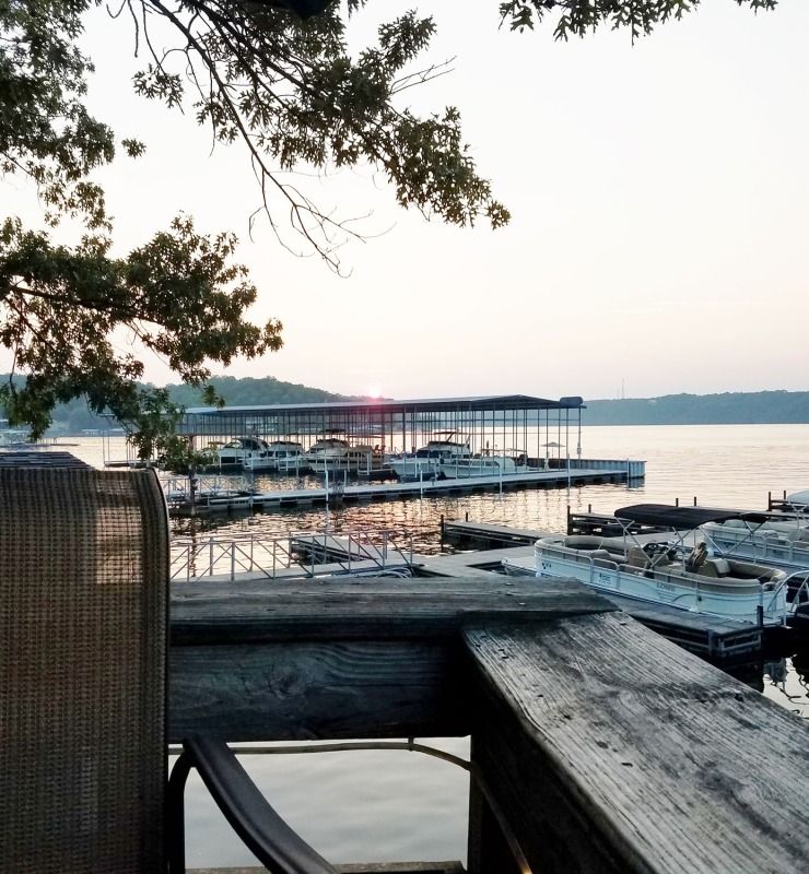 Docked boats at a calm marina beside a lake at sunset, framed by tree branches and a wooden railing