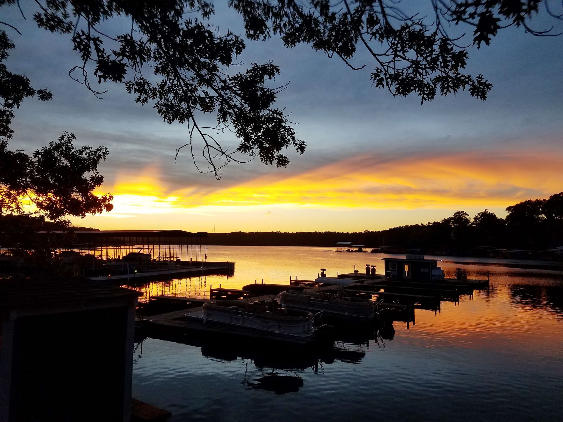 Sunset over a calm marina, with silhouetted trees, docks, and boats reflecting orange light on the water.
