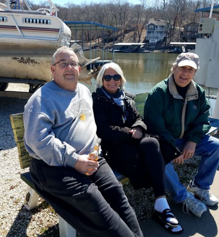 Three people sitting outdoors by boats at a marina on a sunny day.