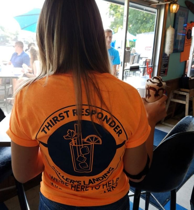 Woman in orange “Thirst Responder” shirt seated in a casual bar or cafe, seen from behind.