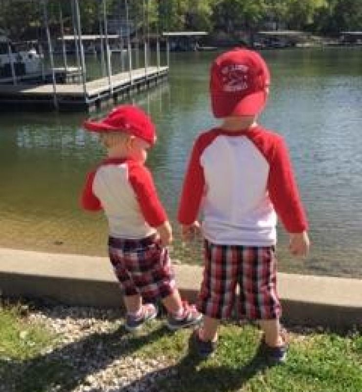 Two children in matching red shirts and plaid shorts standing by a lakeside dock