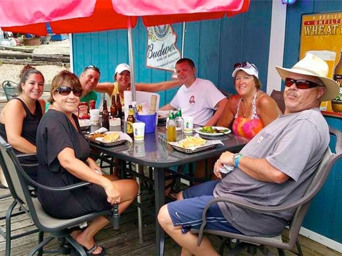 People seated at an outdoor table under a red umbrella beside a blue building, eating and chatting