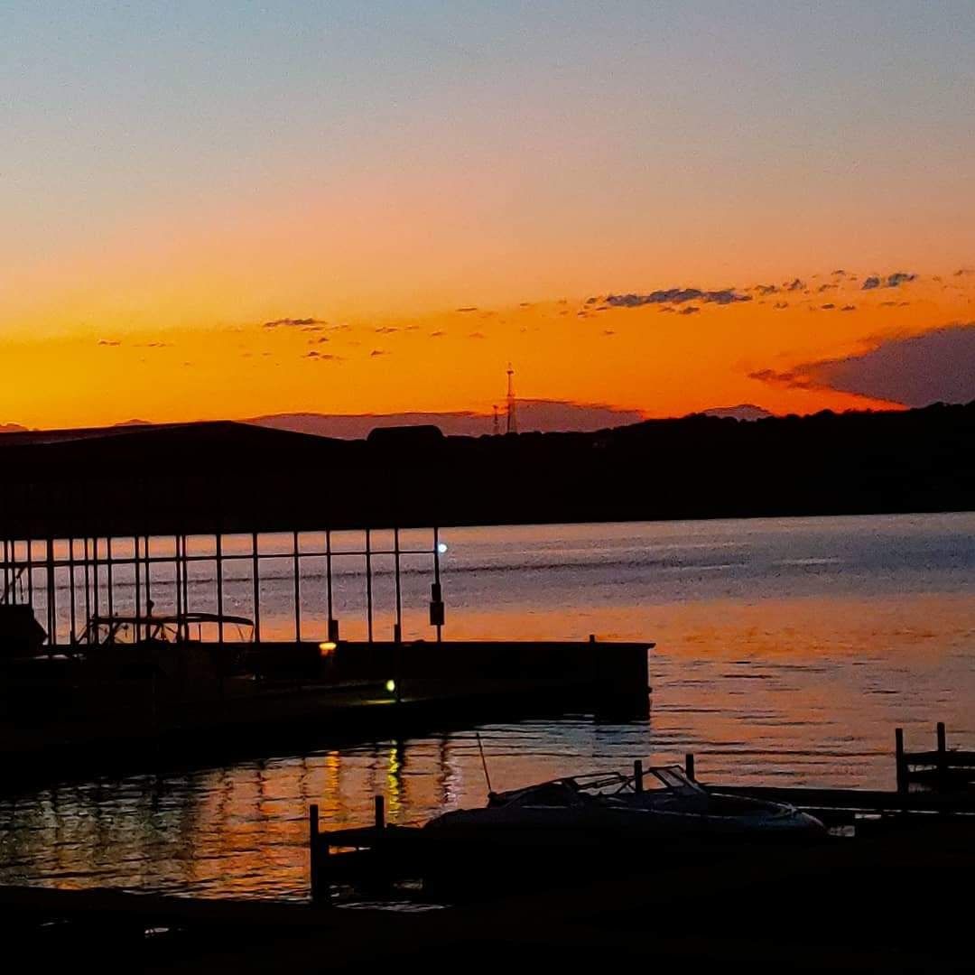 Sunset over a calm waterfront with silhouetted docks, mountains, and orange sky reflected on the water