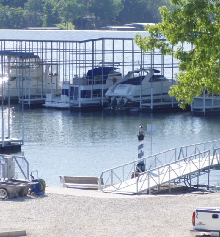 Covered boat slips by a calm lakeshore, with sailboats docked behind a metal railing and a white ramp in front