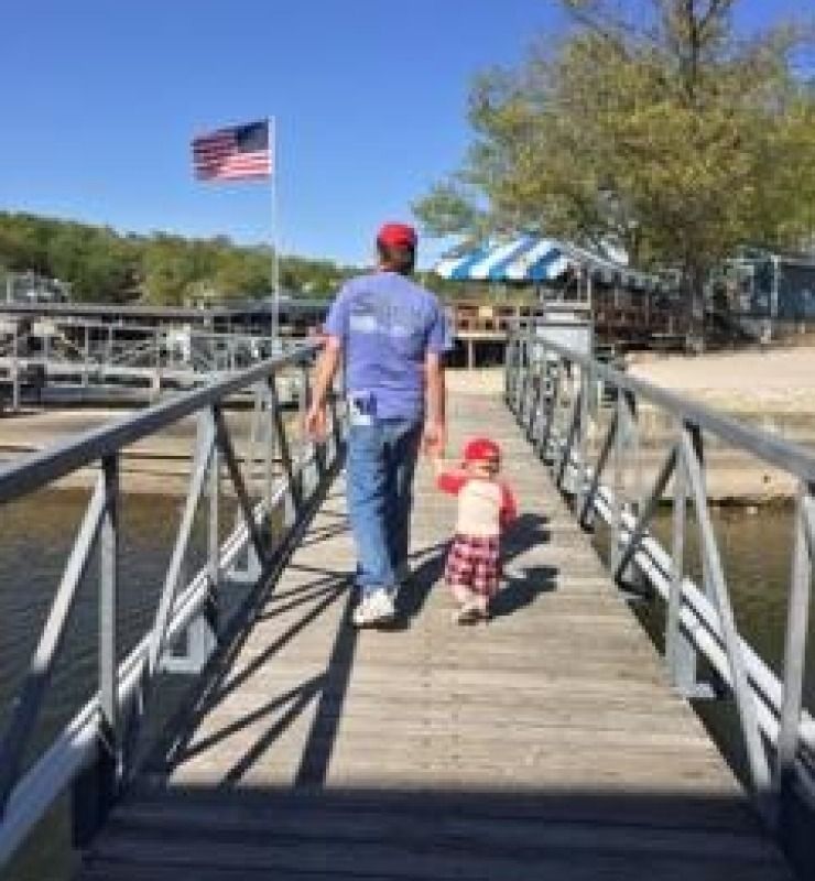 Adult and toddler walking on a wooden dock beside the water, with an American flag in the background