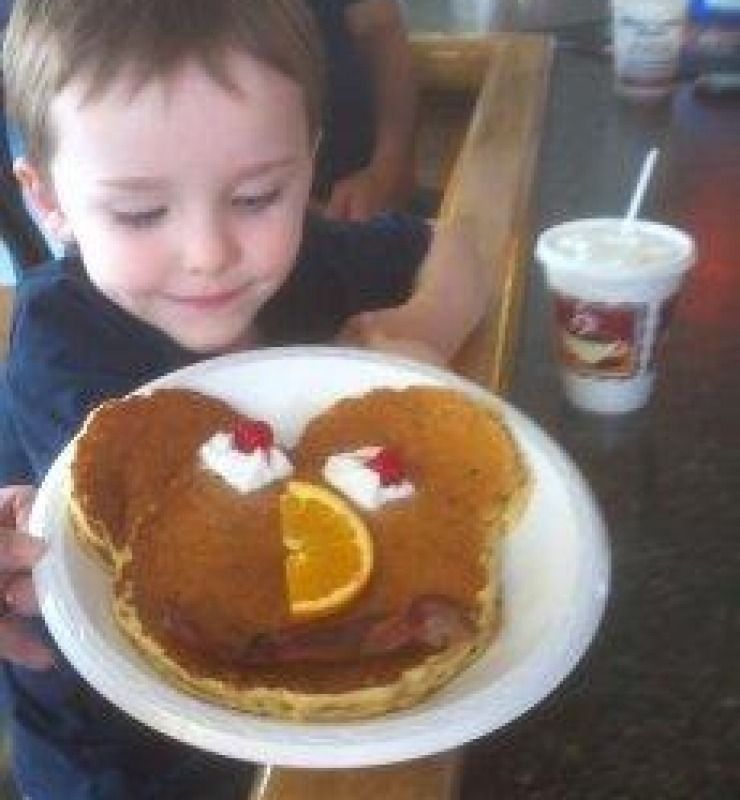 Child smiling at a plate of heart-shaped pancakes with orange slices and whipped cream in a café