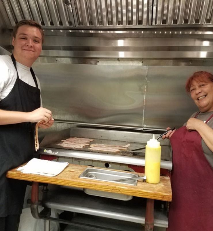Two people smiling beside a grill in a commercial kitchen, holding a tray and squeeze bottle.