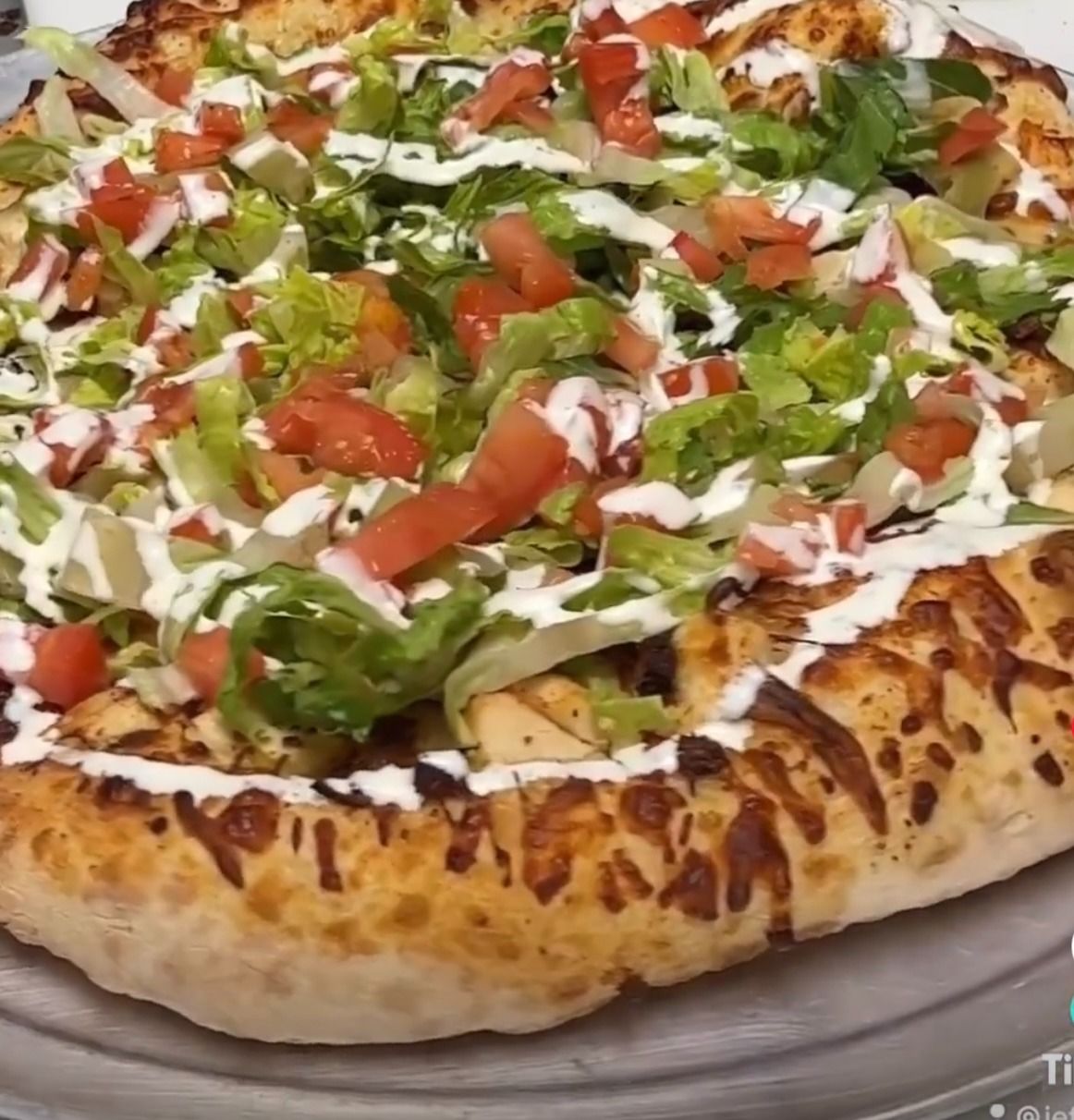 Large loaded pizza with browned crust, topped with lettuce, tomatoes, and drizzled white sauce.