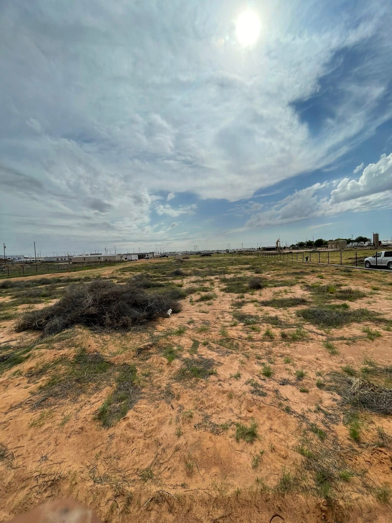 Sandy open field under a bright cloudy sky, with sparse grass and distant structures.