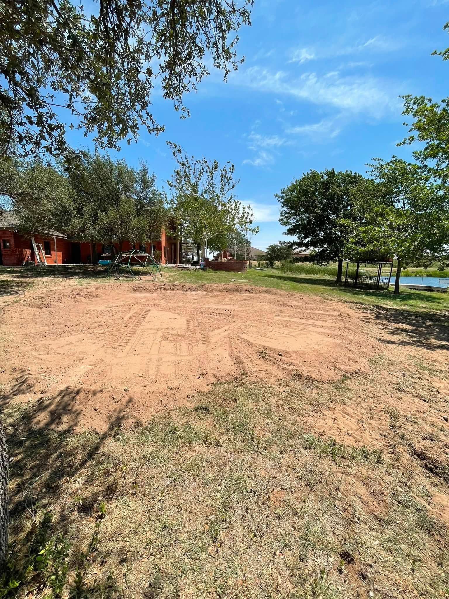 Dirt clearing beside trees under a blue sky, with a road and water in the distance.