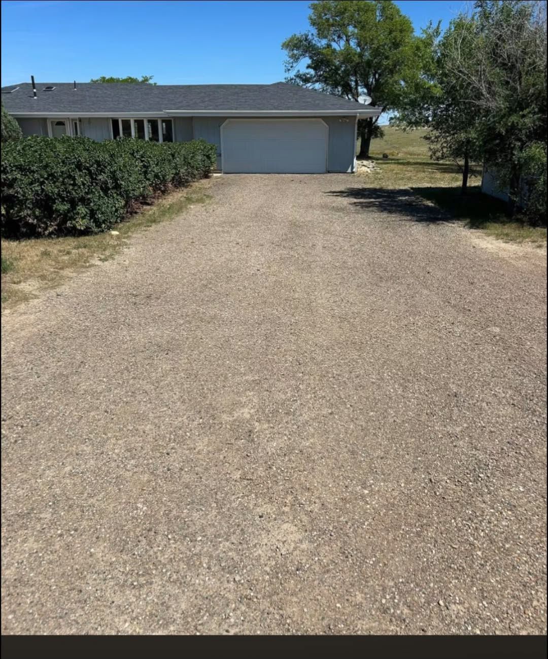 Gravel driveway leading to a single-story house with a garage, shrubs on the left, and trees on the right.