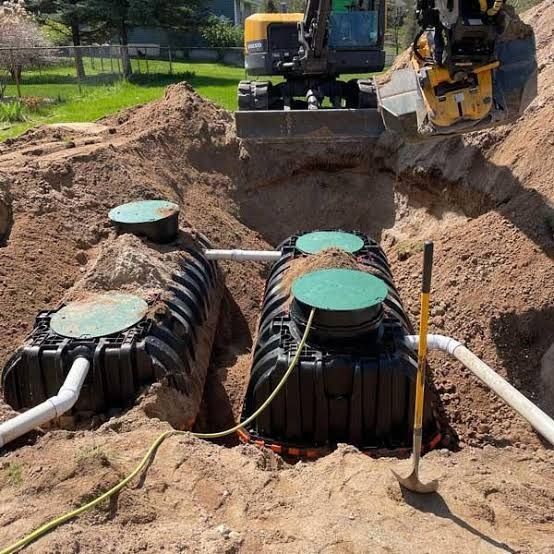 Construction trench with two large black septic tanks, an excavator, and a shovel on a sunny worksite.