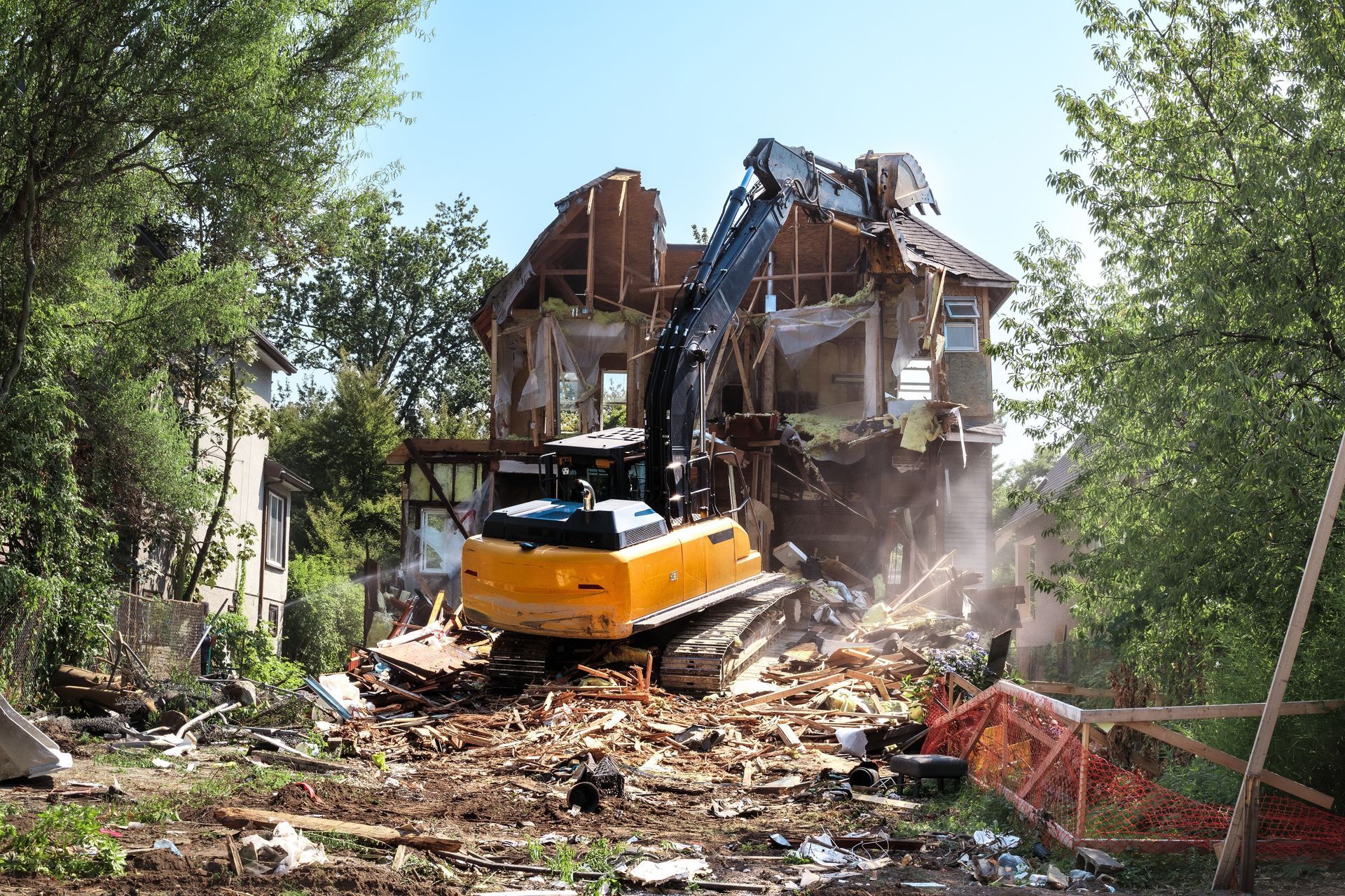 Excavator demolishing a house, with rubble and dust in a tree-lined residential lot.