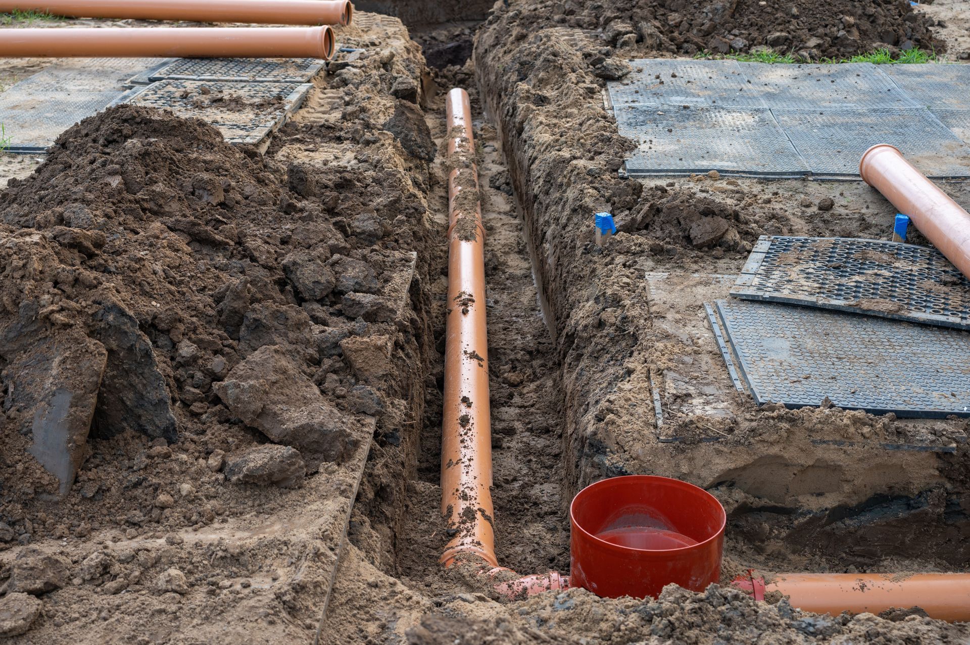 Trenches with orange PVC pipes at a construction site, with a red bucket and dirt excavation.