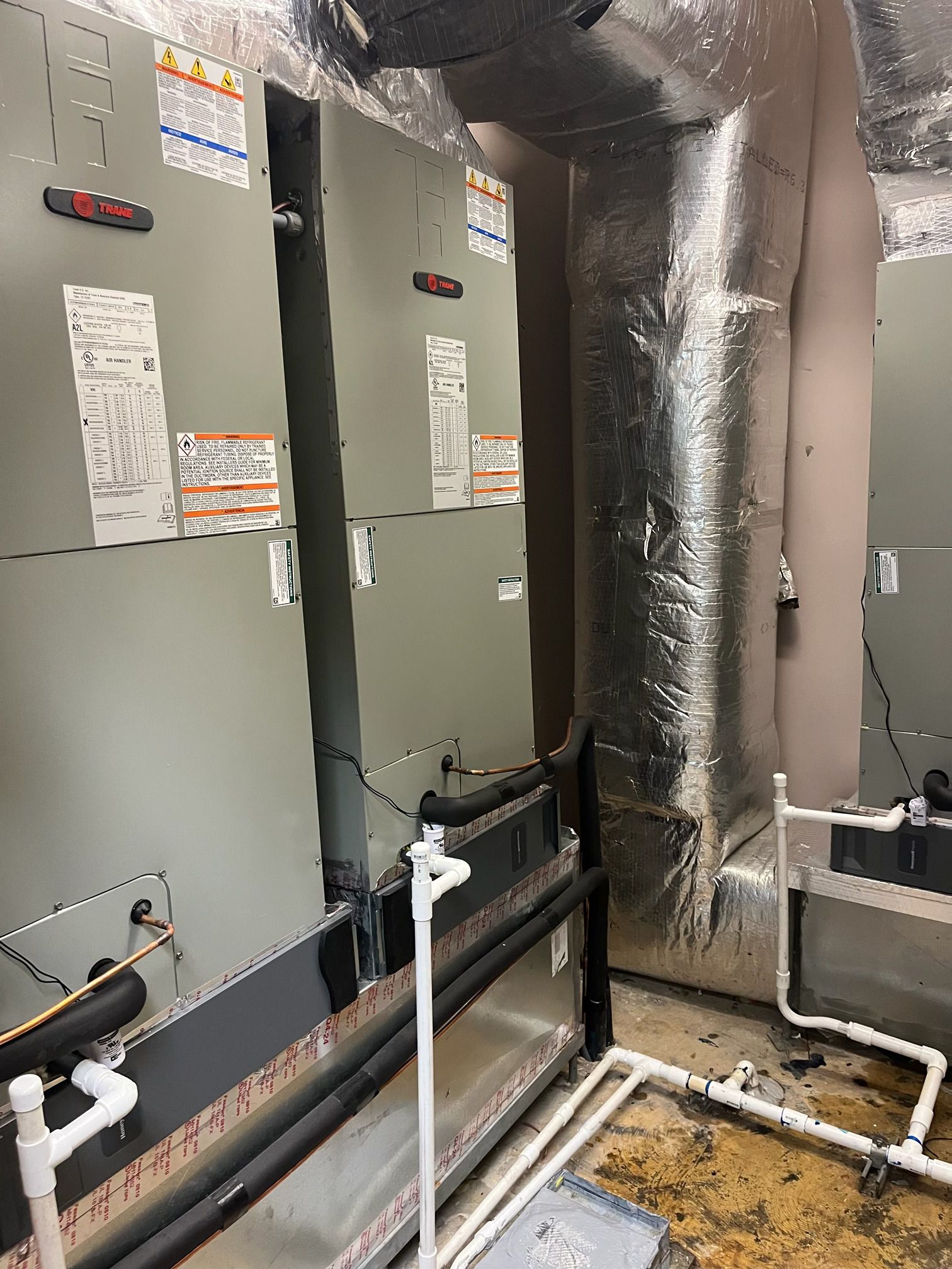 Two gray HVAC air handler units standing side-by-side with connected white PVC drainage pipes in a utility space.