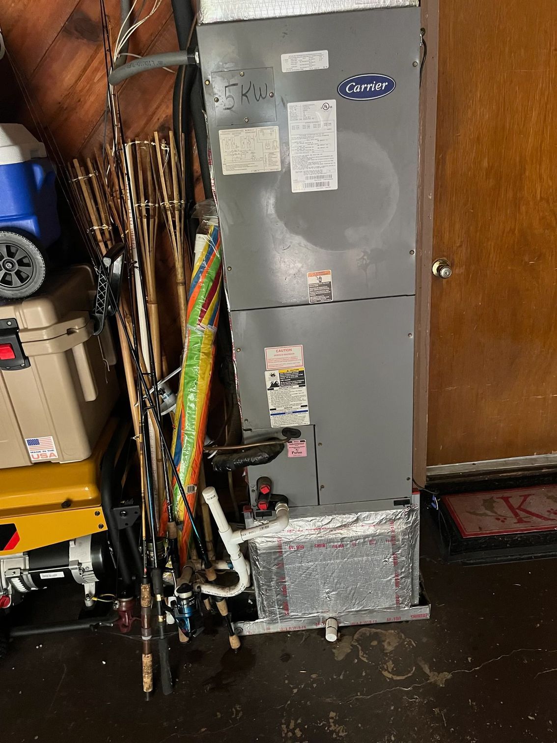 A gray Carrier HVAC unit stands in a room beside a collection of fishing rods and a tan cooler.