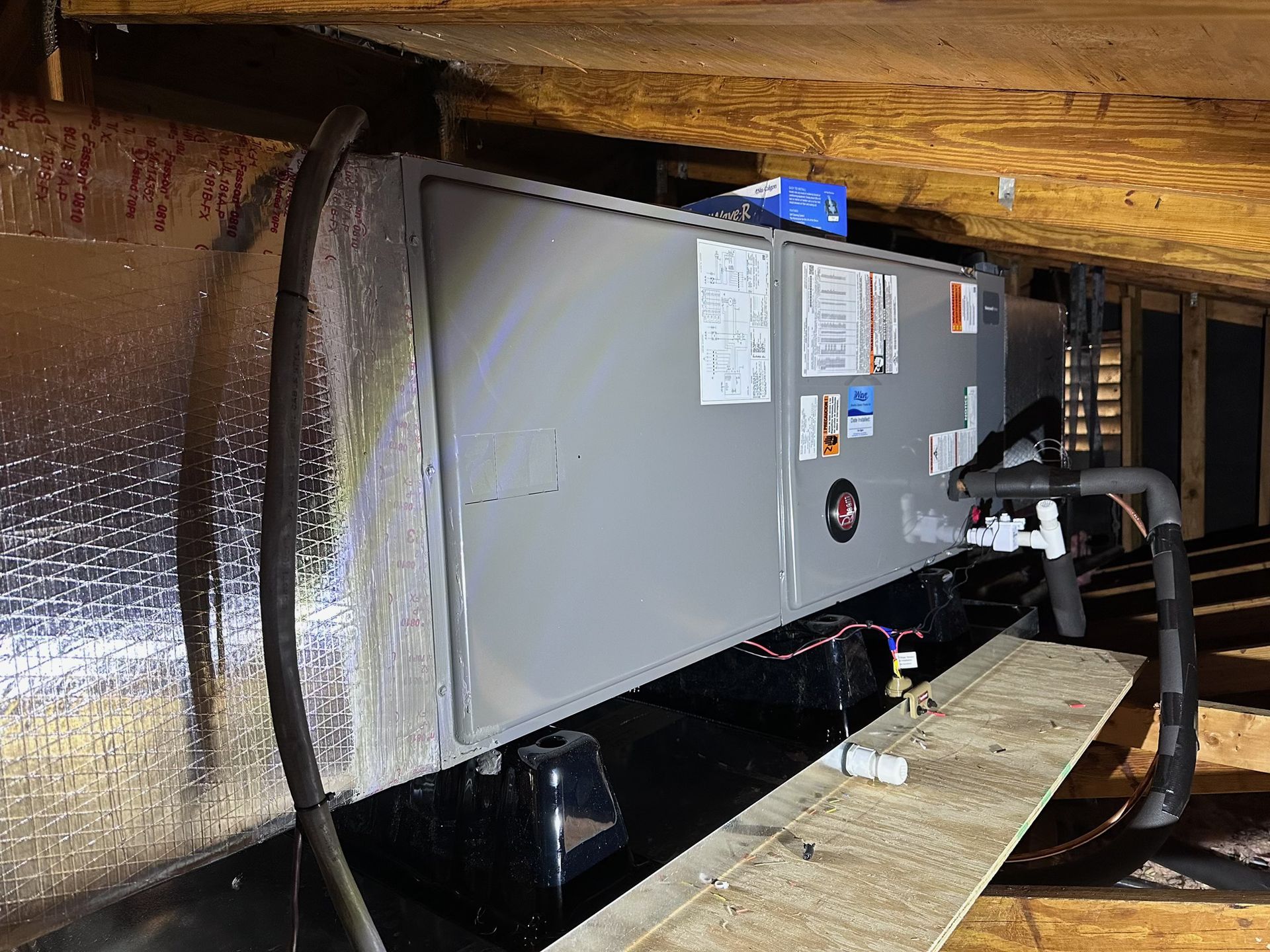 An HVAC air handler unit installed in an attic crawlspace, featuring silver metal casing and insulated piping.