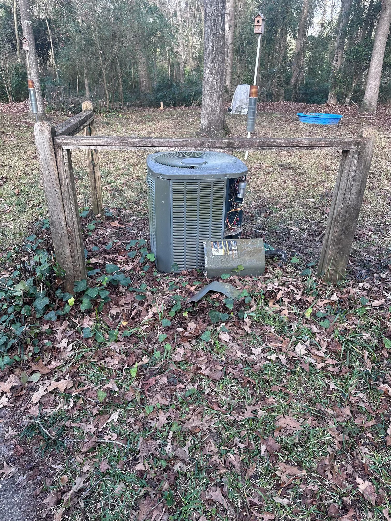 An outdoor HVAC unit sitting on the ground inside a wooden frame in a yard covered in fallen leaves.