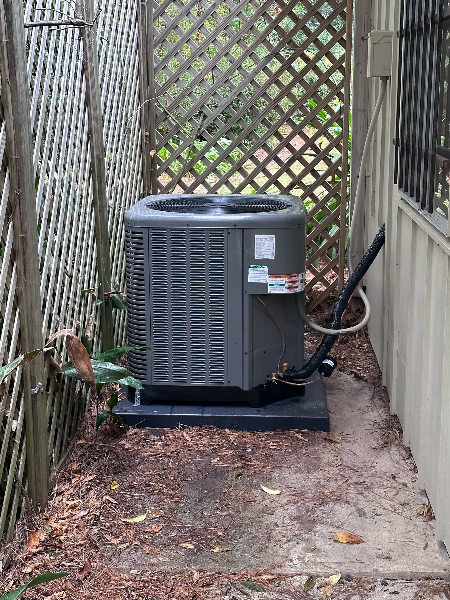A gray air conditioning condenser unit sits on a black pad between a lattice fence and a building exterior.