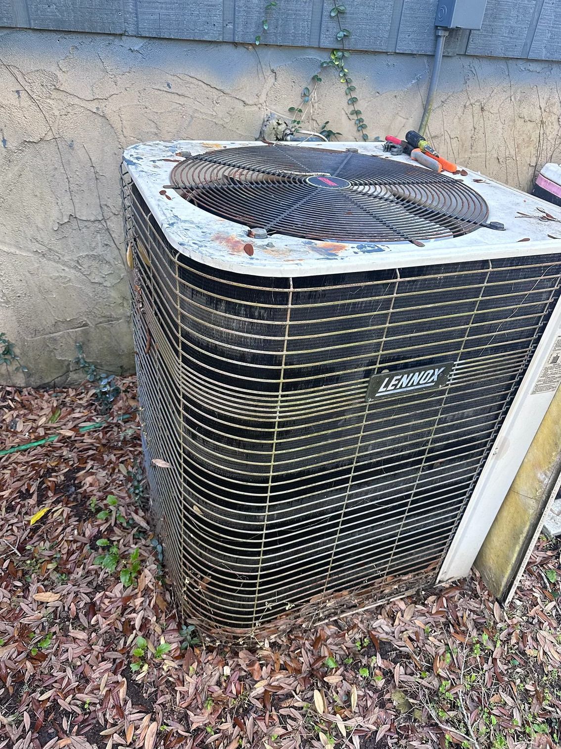 An outdoor air conditioning condenser unit sitting on fallen leaves next to a textured beige wall.