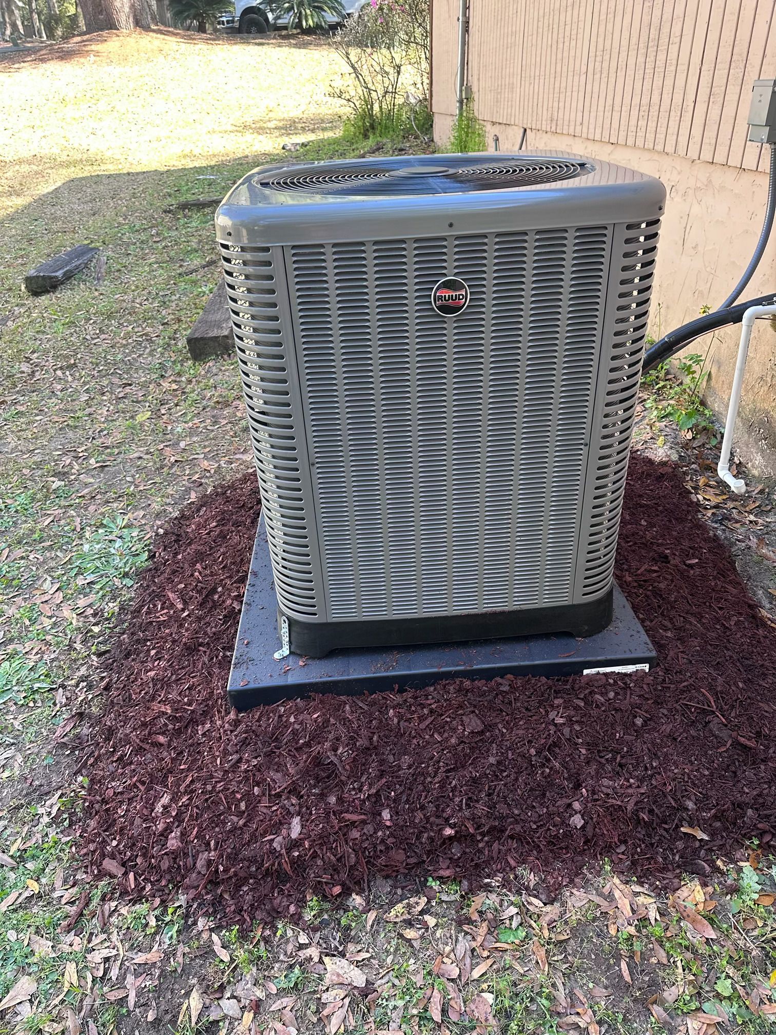A tan outdoor HVAC condenser unit sits on a black pad surrounded by dark mulch against a beige building exterior.