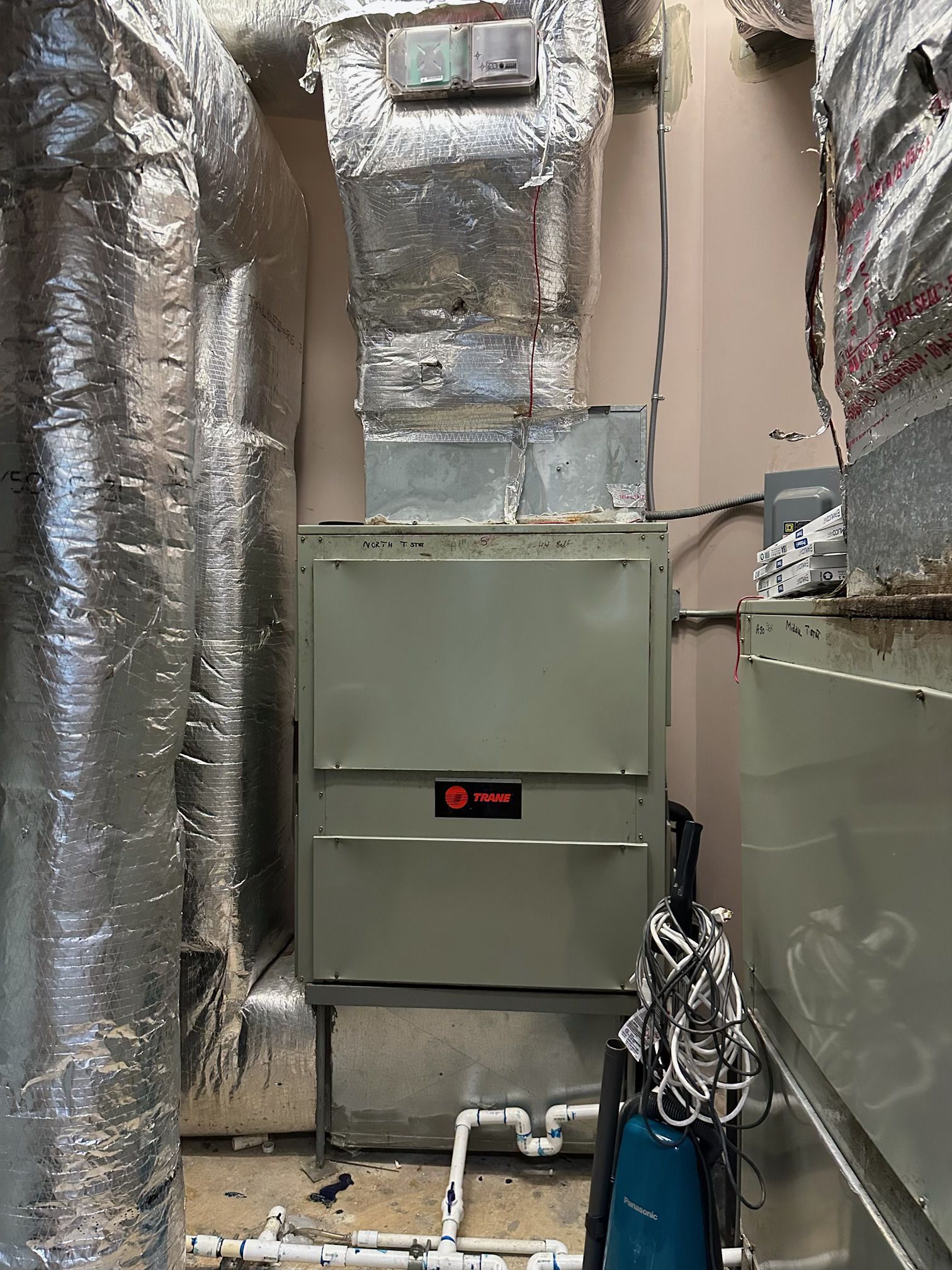 A Trane HVAC unit sits in a utility closet with insulated silver ductwork, white PVC drainage pipes, and a pump.