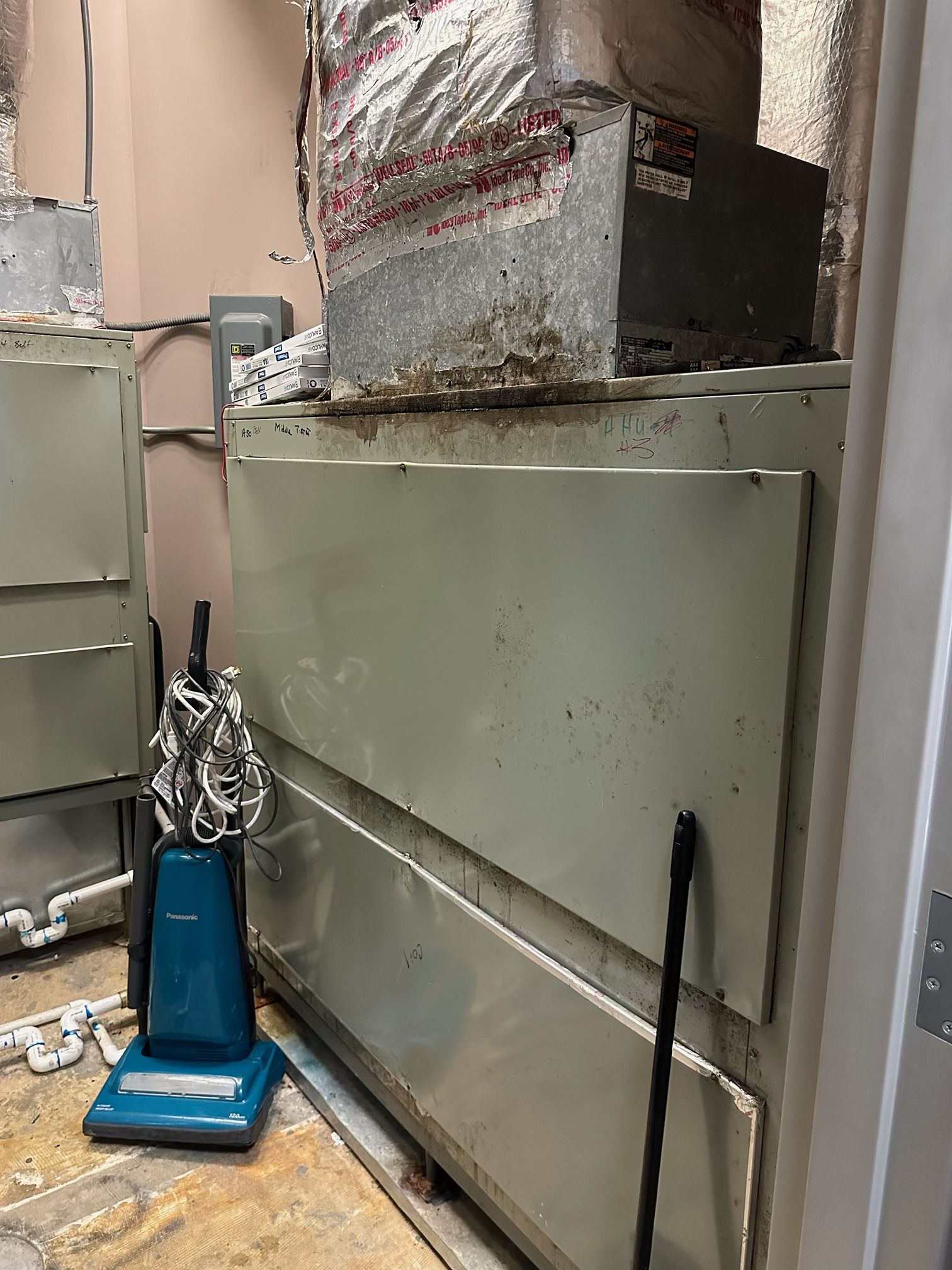 A large, light-green industrial HVAC unit in a utility room, with a vacuum cleaner parked beside it.
