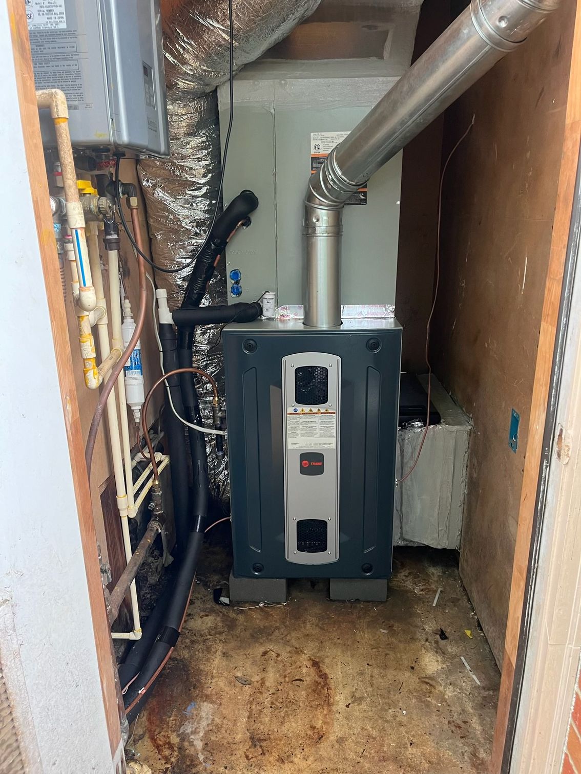 A Trane HVAC furnace unit sits in a utility closet with exposed metal venting and plumbing against wood-paneled walls.