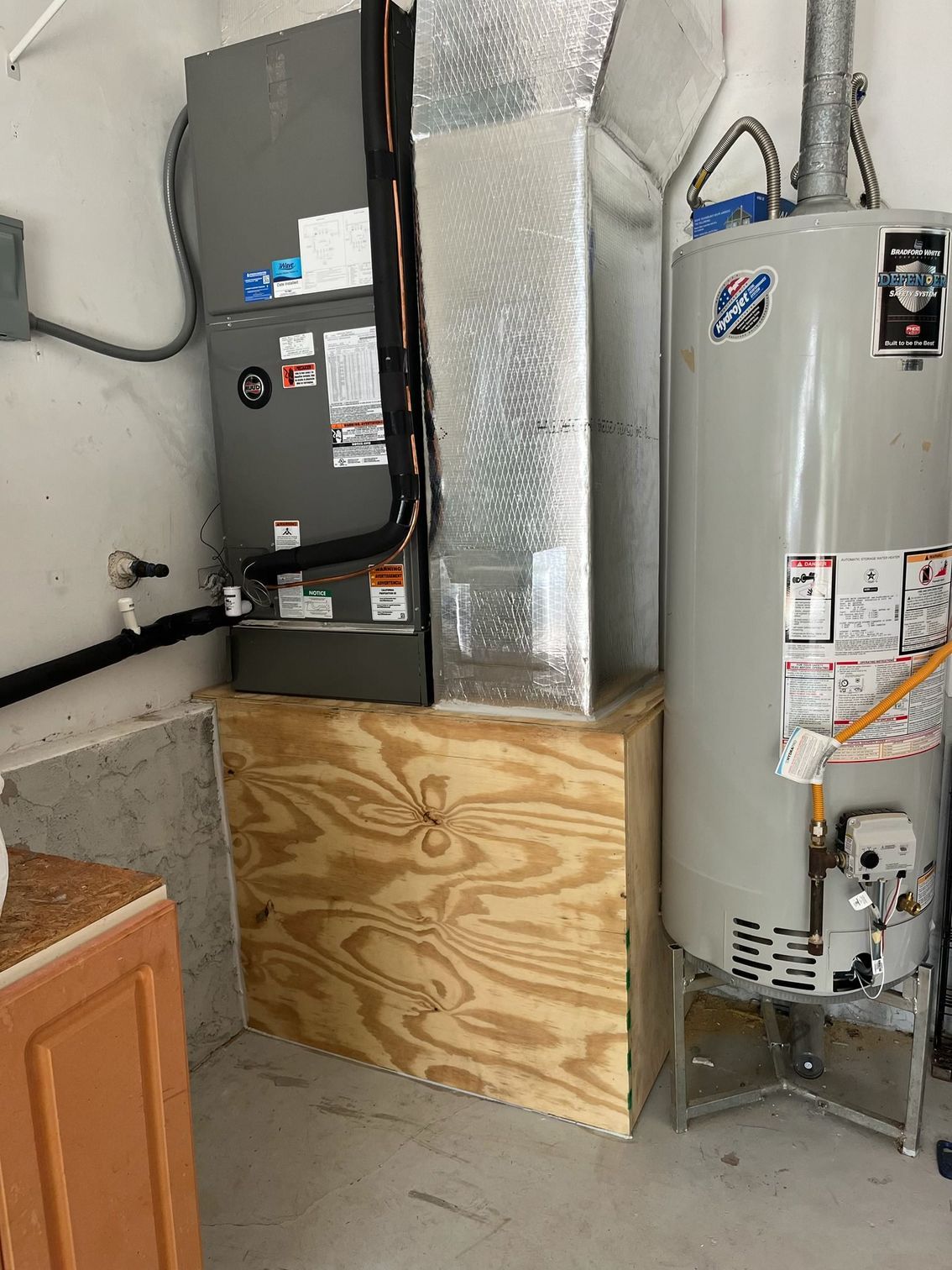 A gray HVAC furnace unit sits on a wooden platform next to a tall, cylindrical gas water heater in a room corner.
