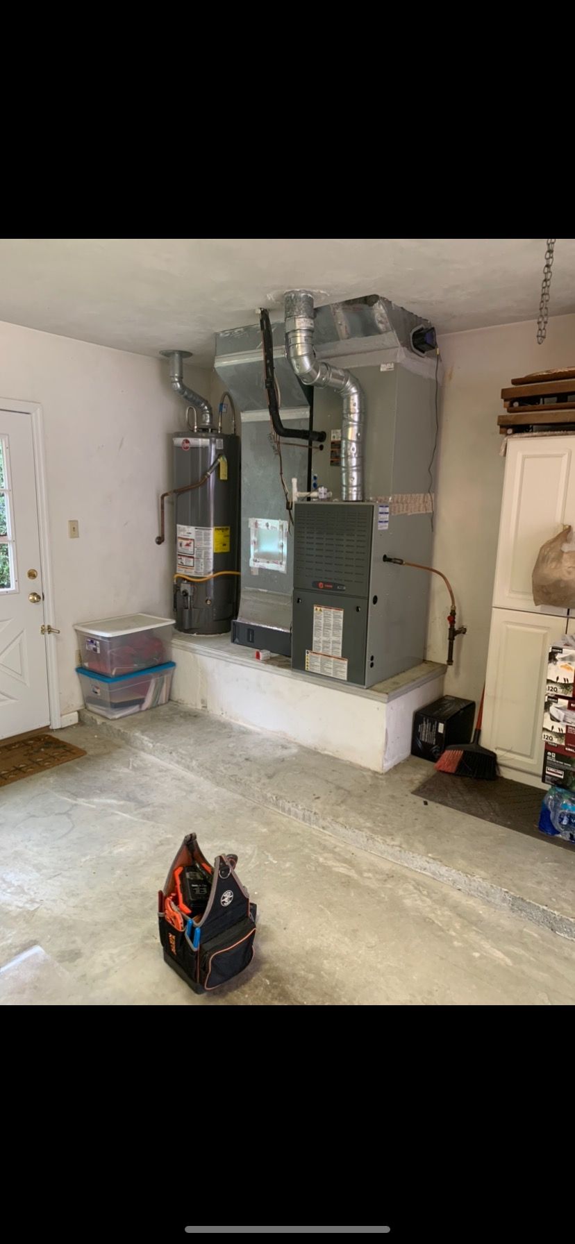 A water heater and a grey HVAC furnace unit sit on a raised concrete platform in a garage near a door and tool bag.