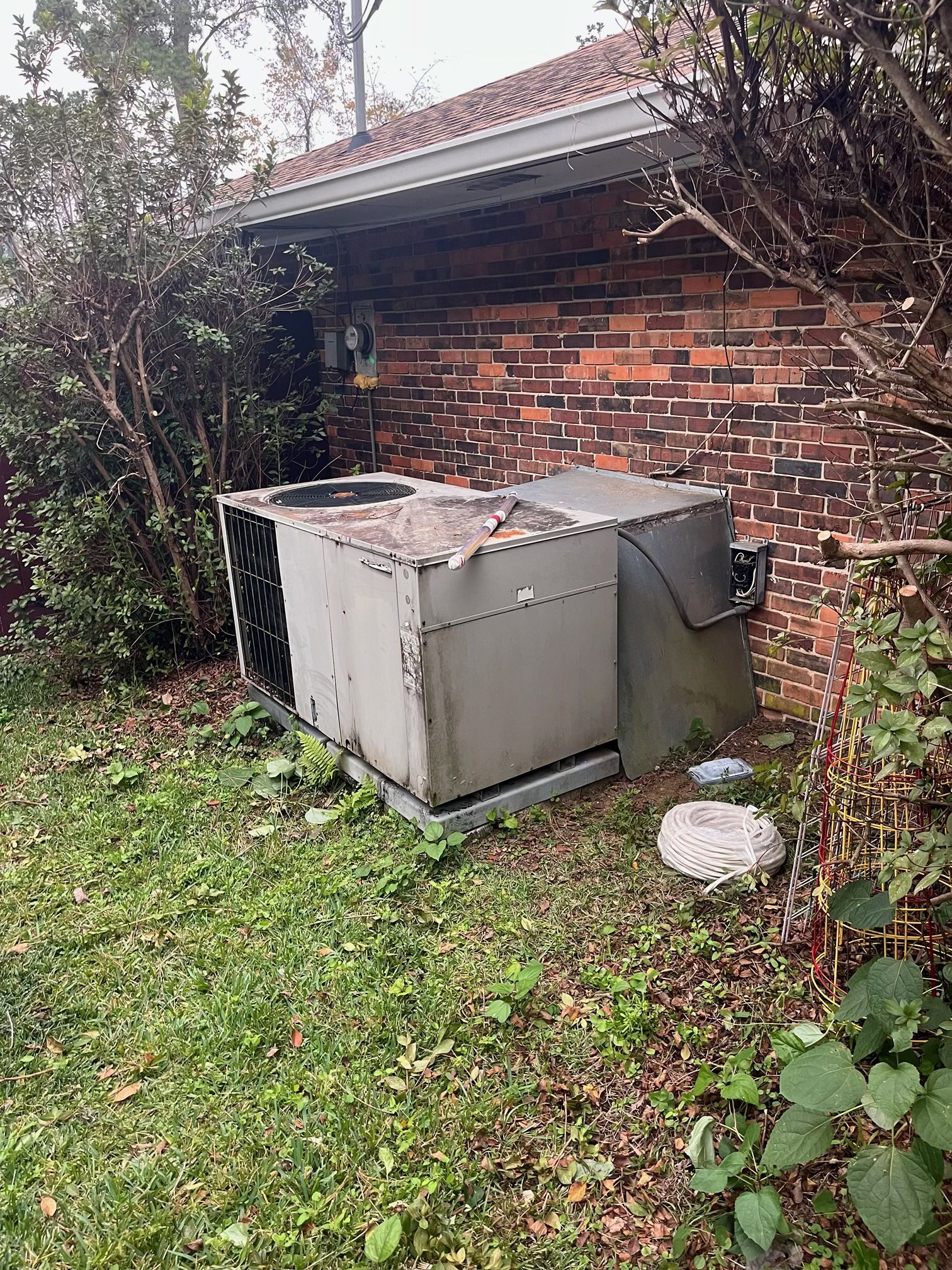 A metal HVAC unit sits on a concrete pad against a brick house wall, surrounded by overgrown grass and bushes.