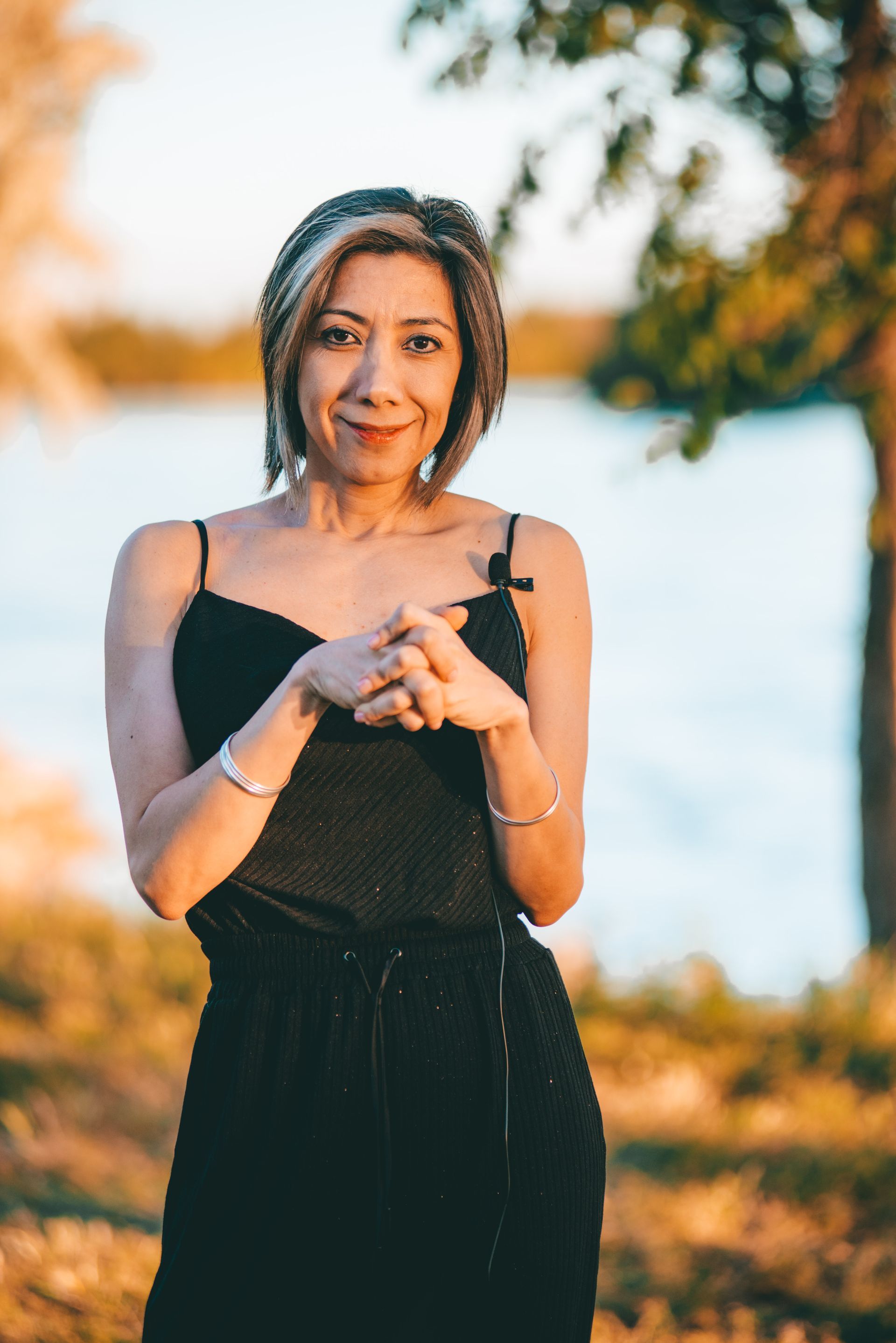 Mujer con mono negro, con las manos entrelazadas, sonriendo cerca del agua con árboles al fondo.