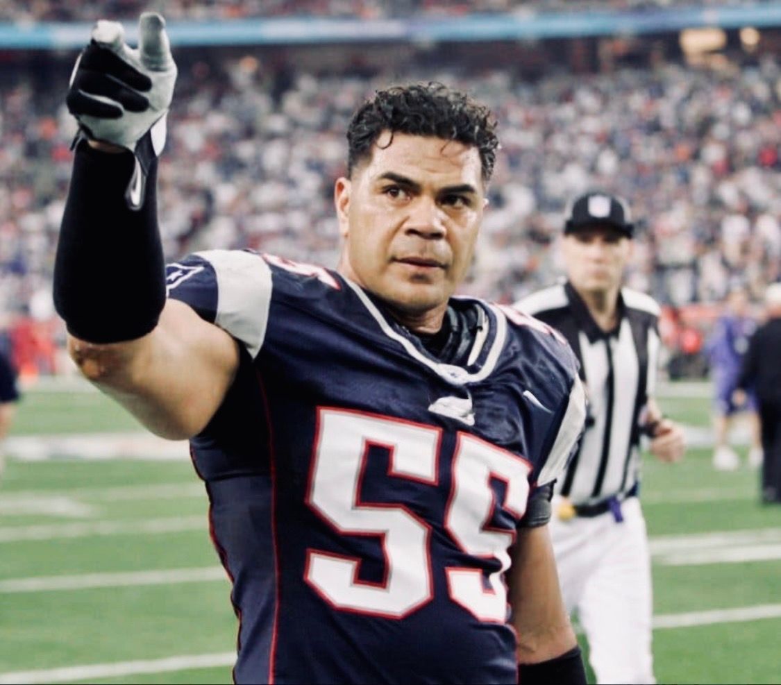 Football player in a navy jersey with number 55 gestures on the field, with a referee and crowd behind him.