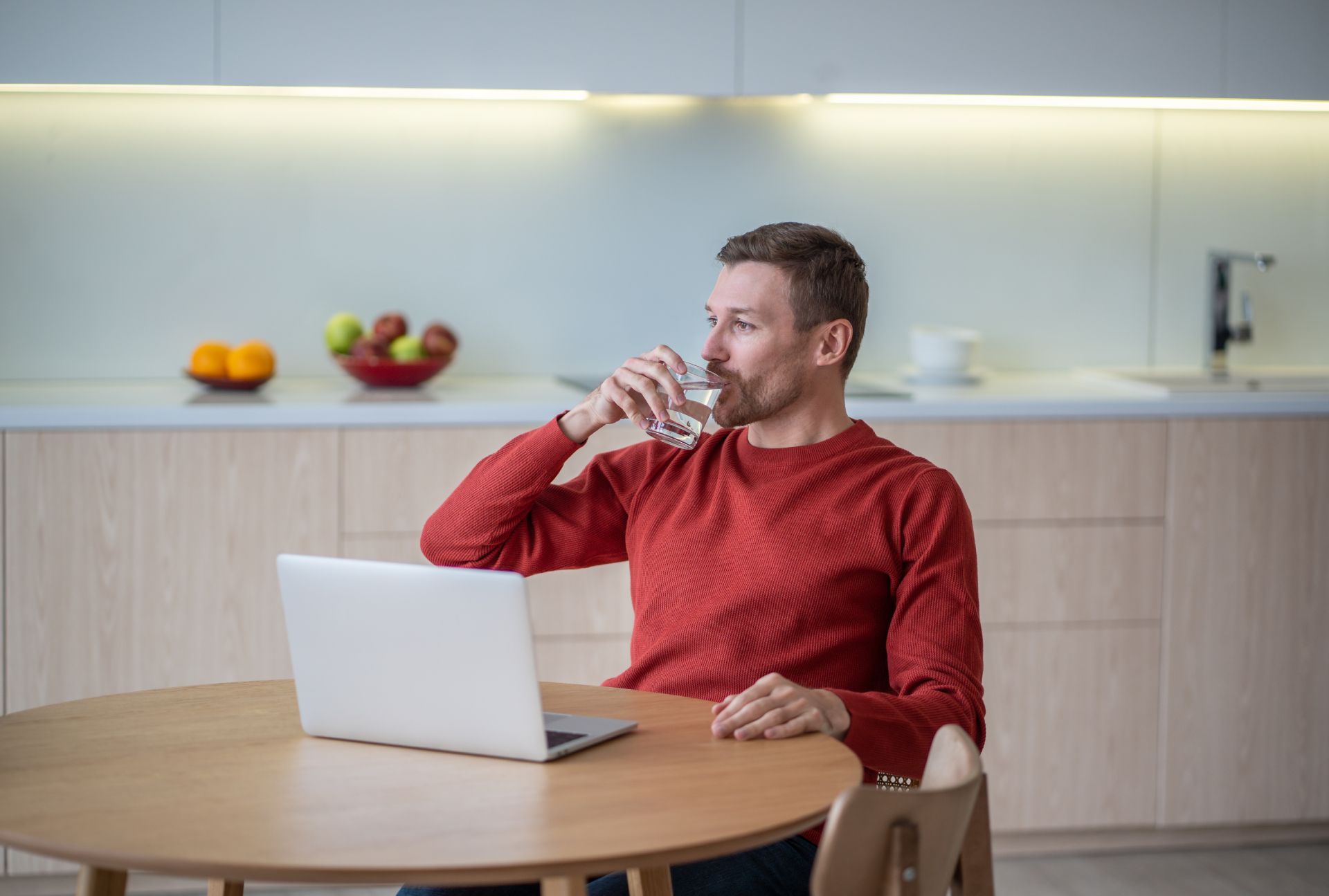 Man in red sweater drinking water at a dining table with an open laptop in a modern kitchen
