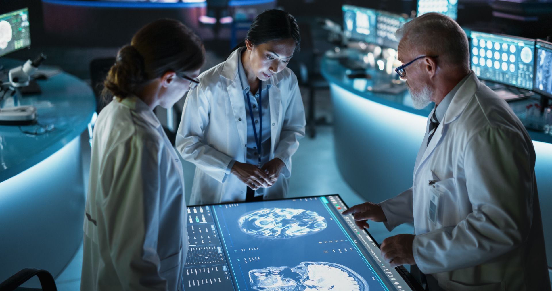 Three lab technicians in white coats review glowing blue data at a high-tech workstation.