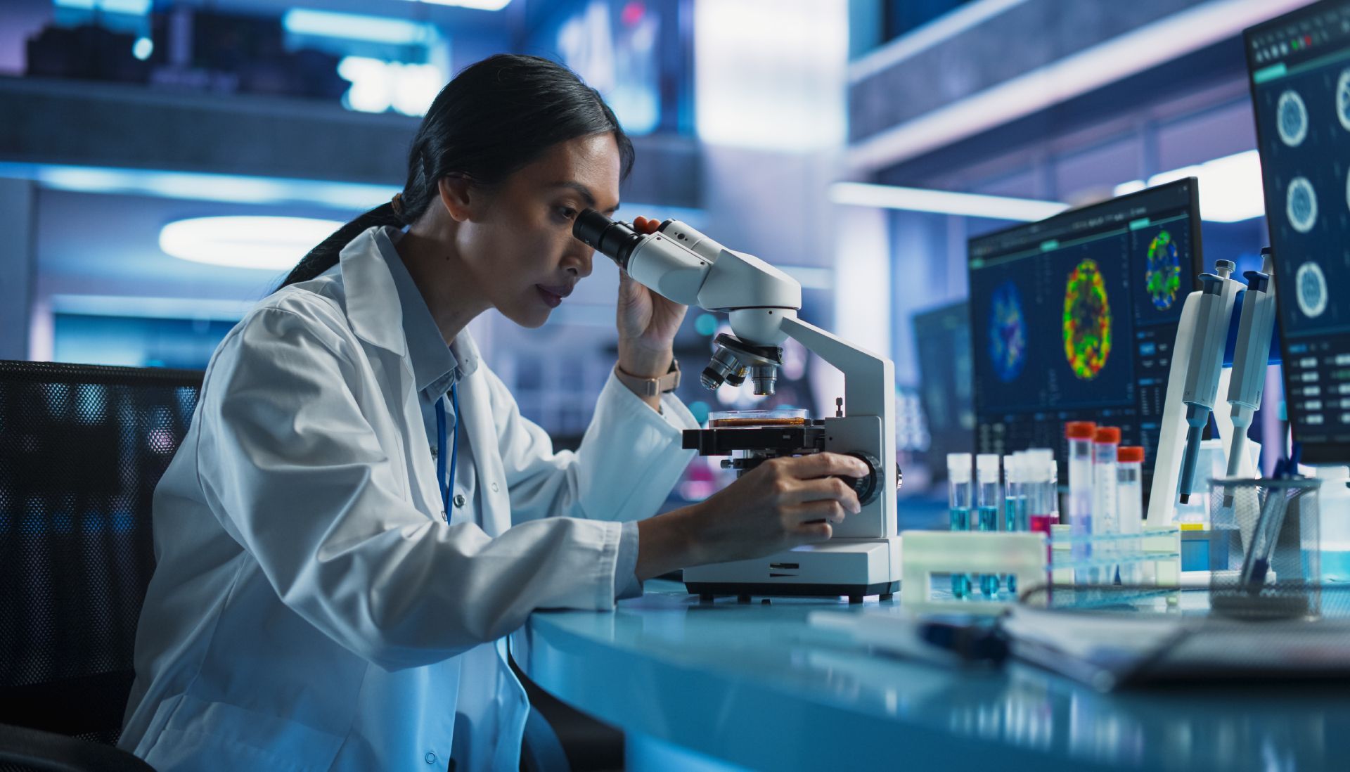 Scientist in a lab coat using a microscope in a modern laboratory with monitors and glassware.