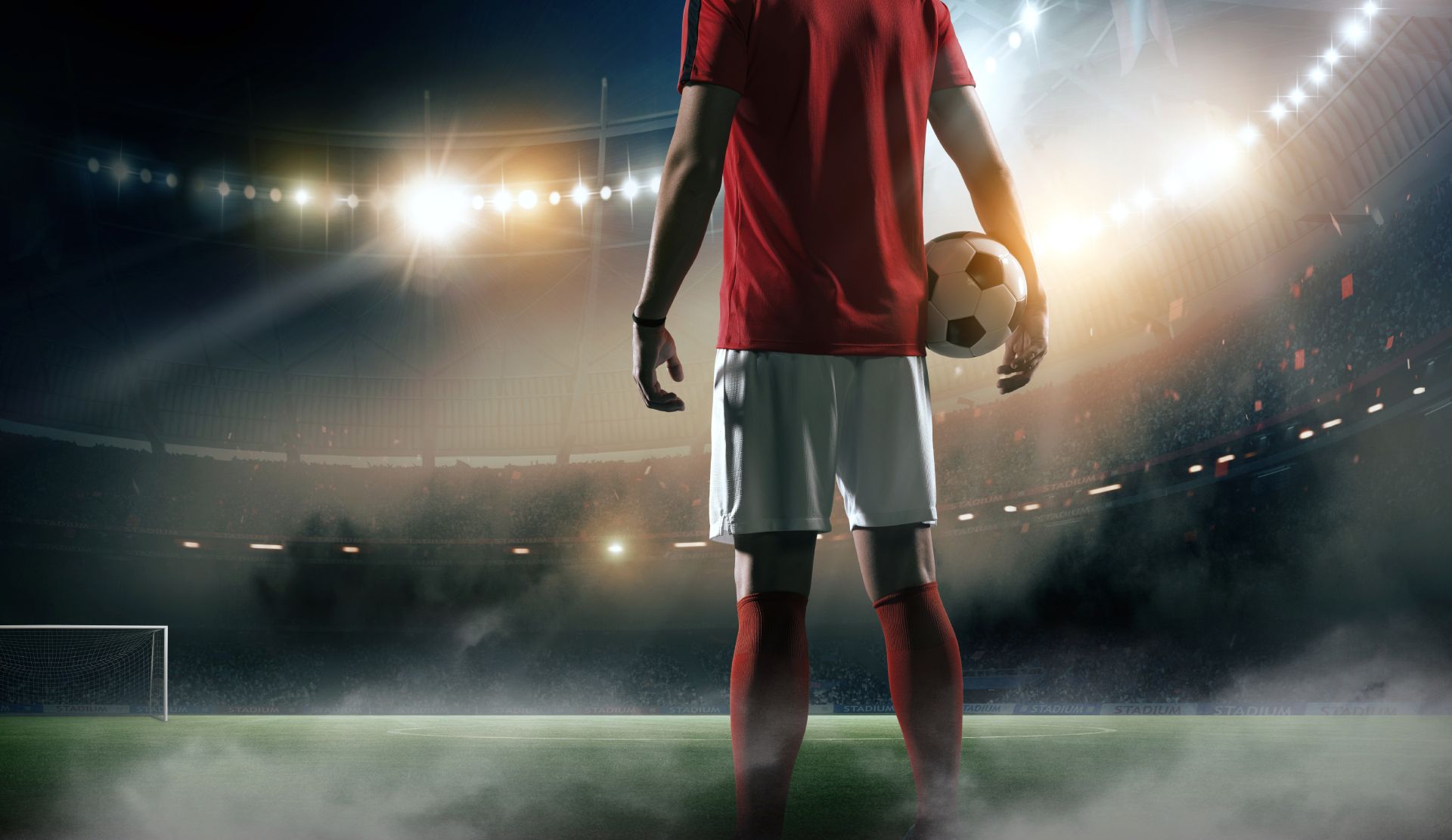 Soccer player in red jersey standing on a lit field, holding a ball before a goal