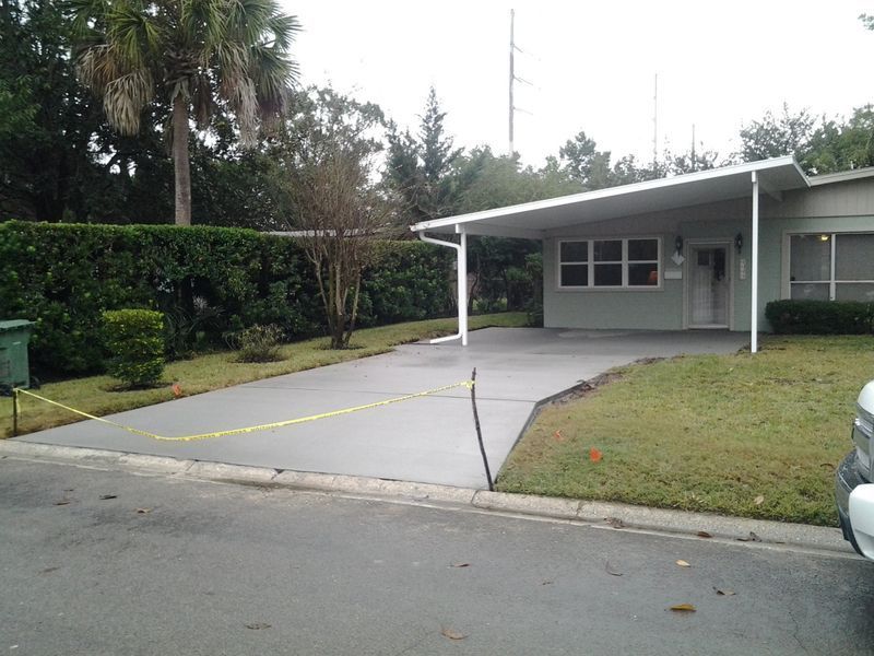 Driveway leading to a light green house with a carport. Lawn and hedges are visible.