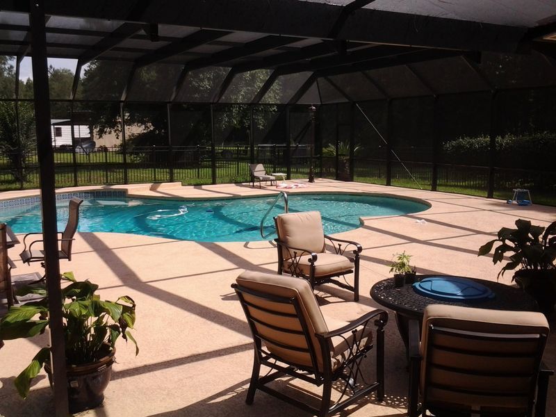 Pool enclosed in a screen, with chairs and plants on the deck. Bright blue water. Sunny outdoor setting.