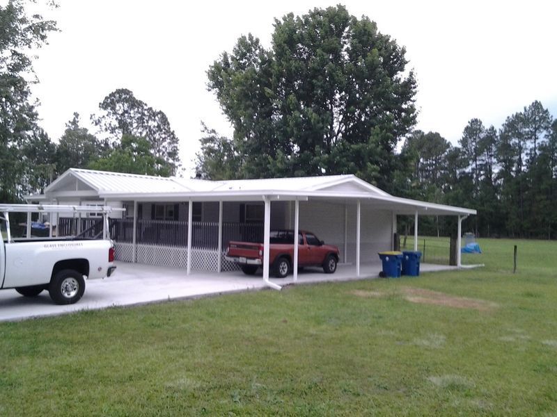 White house with carport, two trucks parked. Green lawn, trees in background. Cloudy day.