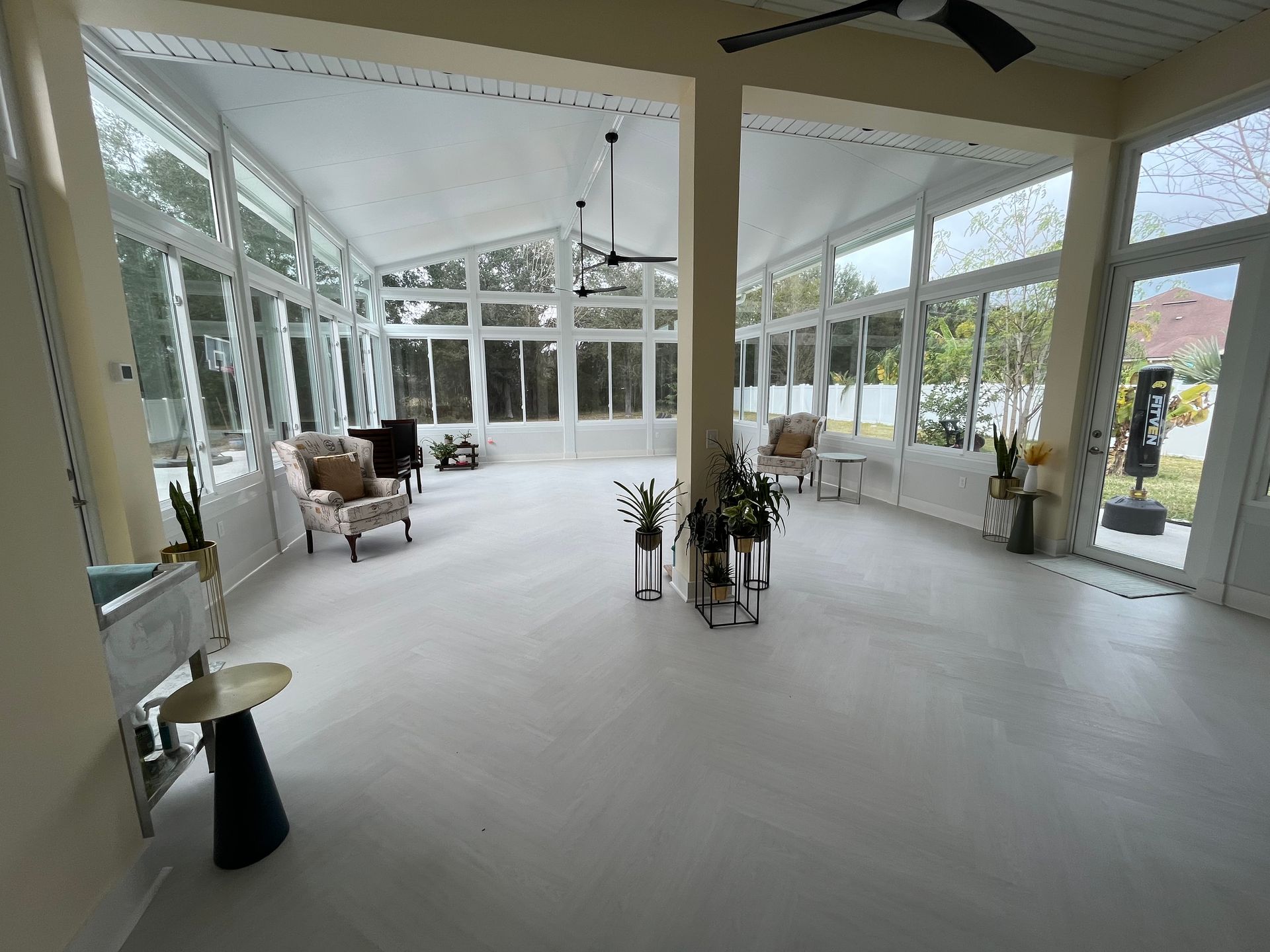 Sunroom with white walls and a light floor, multiple windows, two chairs, plants, and a ceiling fan.