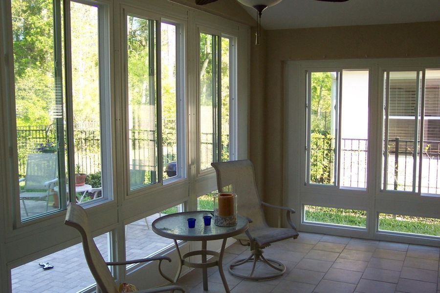Sunroom with white-framed windows and doors; a table, chairs, and view of a balcony and trees.
